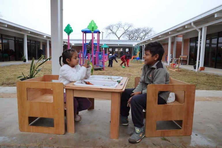 Niña con chaqueta blanca y niño con chaqueta gris sentados en mesa de madera, rodeados de coloridos columpios y otros niños jugando.