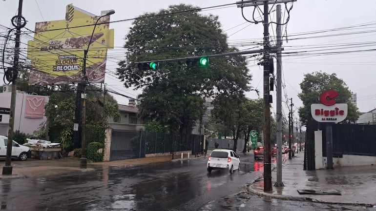 Escena urbana lluviosa en Asunción con un semáforo verde y un cartel de Biggie 24 horas visible.