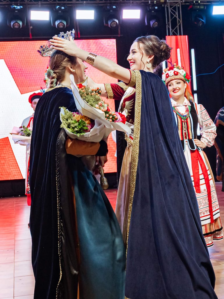 Anabel Sitzmann Hahn en vestido decorado recibe la corona de otra mujer vestida con atuendo tradicional y sosteniendo flores.