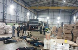 Grupo de detenidos acostados en un almacén, rodeados por agentes de seguridad vigilantes y un vehículo de carga al fondo.