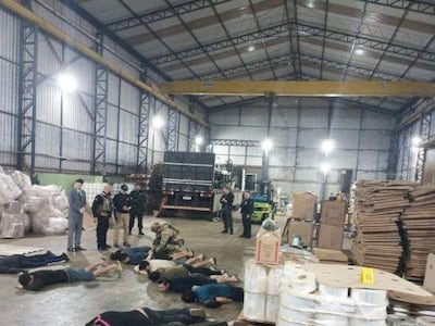 Grupo de detenidos acostados en un almacén, rodeados por agentes de seguridad vigilantes y un vehículo de carga al fondo.