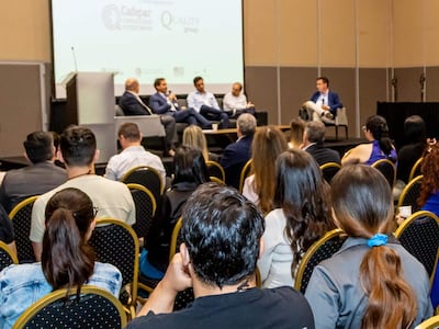 Cinco panelistas sentados, uno hablando mientras el público escucha, todos con camisetas 'BECOME MORE'.