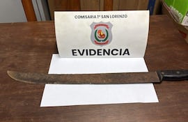 La imagen muestra un objeto cuchillo de gran tamaño, probablemente un machete, que está apoyado sobre una superficie de madera. Al fondo, se observa un cartel que dice "EVIDENCIA" y lleva el emblema de la Policía Nacional de Paraguay, específicamente de la "COMISARIA 1° SAN LORENZO".
No hay personas visibles en la imagen. El ambiente parece ser un espacio interior, posiblemente una oficina o un área de procedimientos policiales, dado el contexto del objeto presentado como evidencia. No hay otros elementos visuales, como equipos de fútbol, ni lugares reconocibles. La acción que se puede inferir es la presentación de este objeto como parte de una investigación o incidente relevante. La imagen se centra en la exhibición del machete y la identificación de su procedencia a través del cartel.