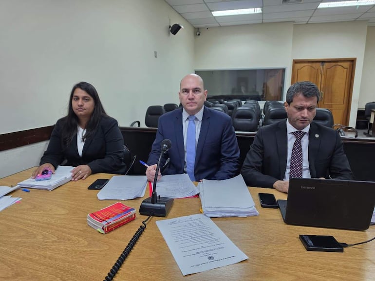 Mujer con saco negro observa mientras abogado en traje azul habla frente a un micrófono en una sala de audiencias.