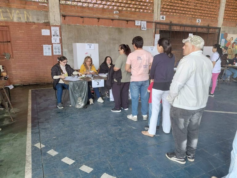 Tres mujeres y un hombre en mesa electoral, con carteles sobre el proceso electoral al fondo.