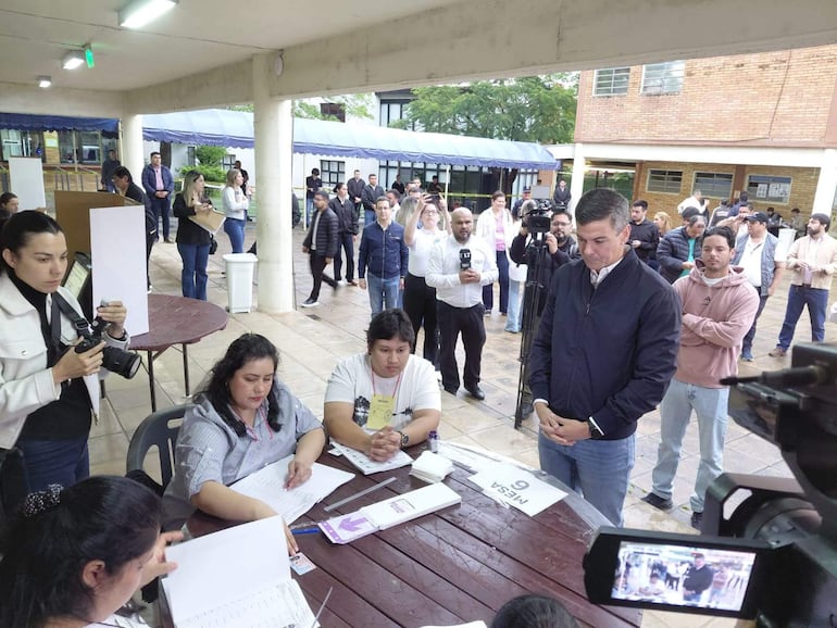 Santiago Peña de pie, vestido oscuro y pensativo, observa la mesa de votación donde una mujer con blusa a rayas registra información.
