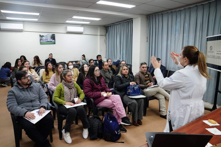 Mujer de bata blanca habla frente a un público atento en salón con cortinas azules, algunos tomando notas.