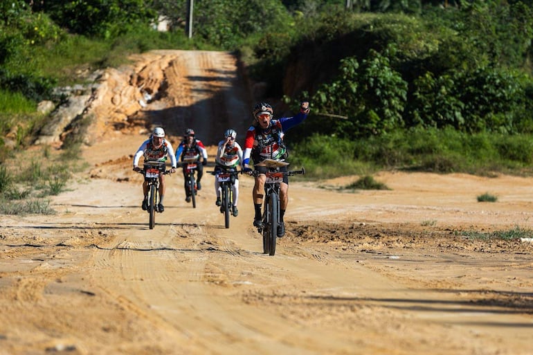 Cinco ciclistas del team Angiru visten camisetas numeradas, uno levanta la mano en señal de triunfo en un camino de tierra.