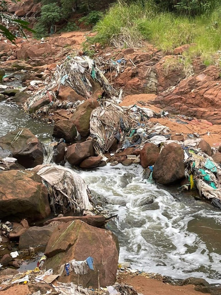 Un curso de agua salpicado de espuma rodeado por grandes rocas y vegetación, con desechos acumulados en la orilla.