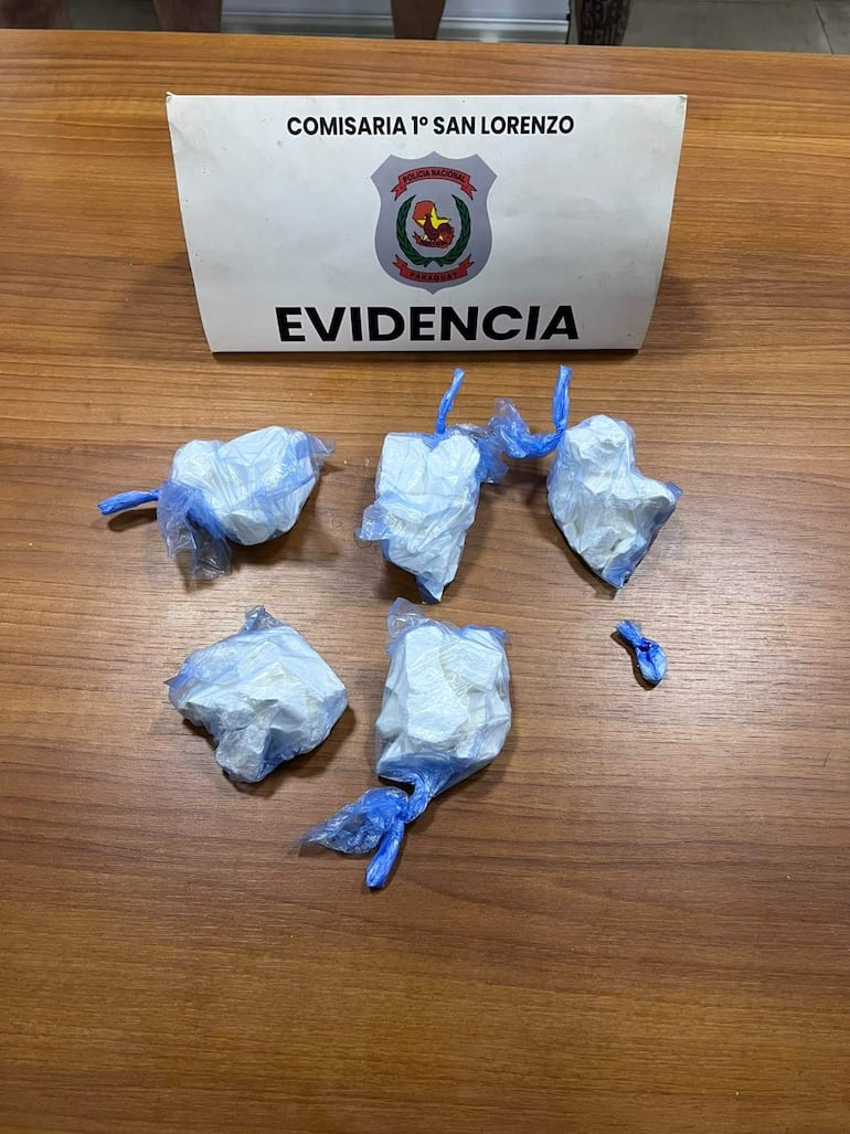 Cinco paquetes envueltos en plástico azul sobre mesa de madera en la Comisaría 1° San Lorenzo, con cartel de evidencia al fondo.