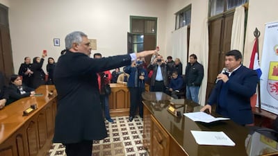 Hombre de cabello gris con chaqueta oscura levanta la mano en juramento, rodeado de personas en un ambiente formal.