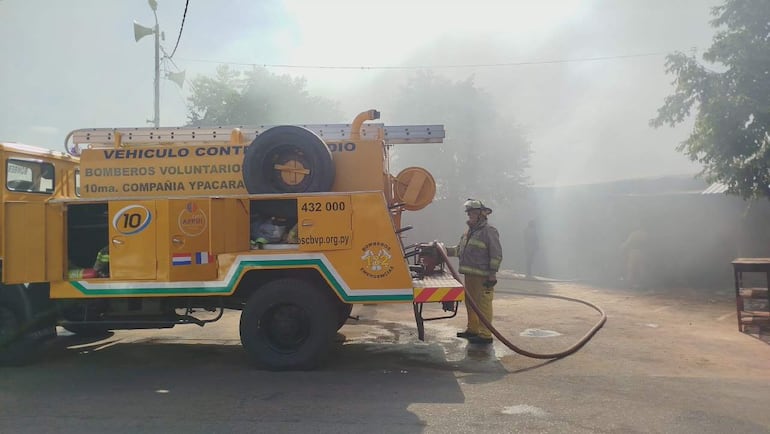 Bombero vestido con equipo de protección sostiene manguera junto a camión amarillo de bomberos en medio del humo.