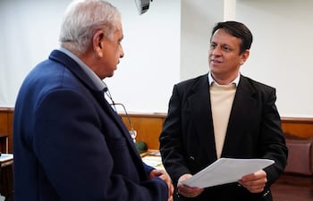 Dos hombres en oficina; uno con chaqueta azul y el otro con ropa oscura sostiene documentos, conversando seriamente.