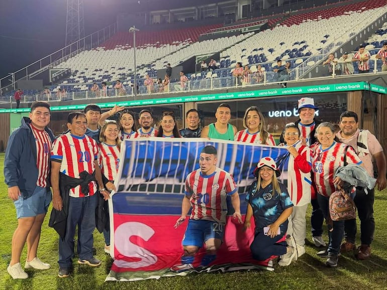 Hinchas de la Albirroja con camisetas rojas y blancas sosteniendo una bandera en un estadio, mostrando expresiones felices.