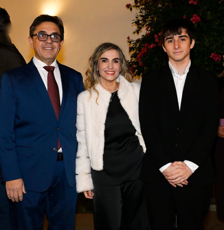 Hombre con traje azul y corbata oscura sonríe junto a una mujer en chaqueta blanca y un joven en traje negro, en ambiente amistoso.