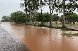 Escena de calle inundada en Concepción, con agua marrón cubriendo la vía y árboles a los lados bajo un ambiente nublado.