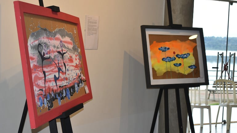 Dos cuadros abstractos en exhibición, uno con marco rojo y otro negro, en galería con vista a un cuerpo de agua.