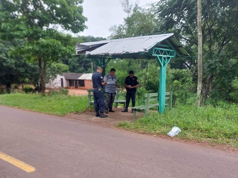 Policías conversan en una parada de bus, uno apartado mirando al suelo. Ambiente rural con vegetación y casas al fondo.