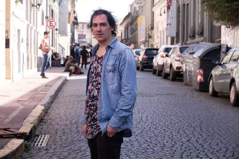 Felipe Barrozo con camisa floral y chaqueta de mezclilla, de pie en una calle tranquila mientras varias personas lo rodean.