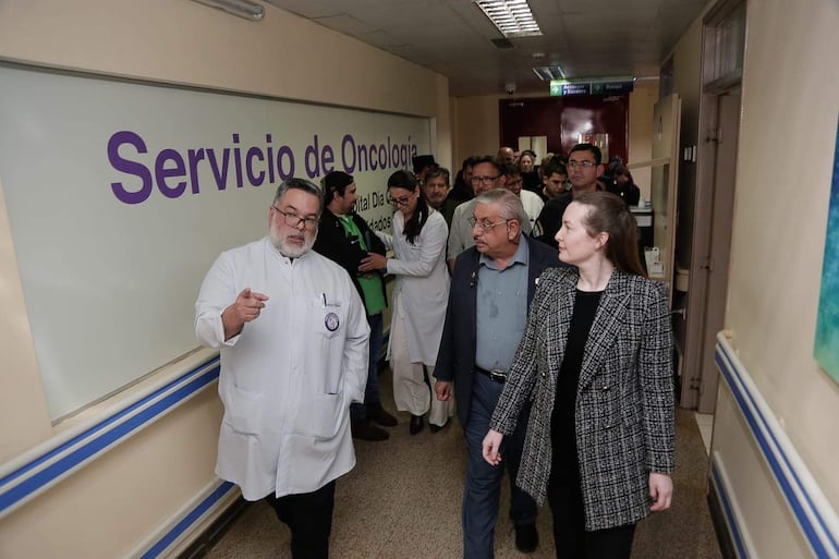 Isaías Fretes, médico de bata blanca y gafas, explica a Derlis León y mujer con abrigo a cuadros en pasillo del hospital.