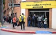 Fila de personas en la vereda, entre ellos una mujer de prenda naranja y un hombre con mochila amarilla y cámara, frente a un edificio.