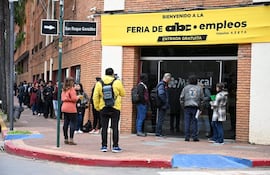 Fila de personas en la vereda, entre ellos una mujer de prenda naranja y un hombre con mochila amarilla y cámara, frente a un edificio.