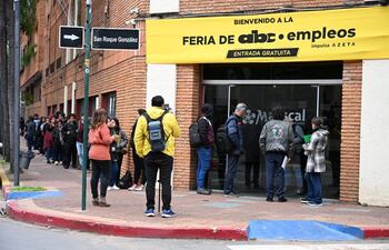 Fila de personas en la vereda, entre ellos una mujer de prenda naranja y un hombre con mochila amarilla y cámara, frente a un edificio.
