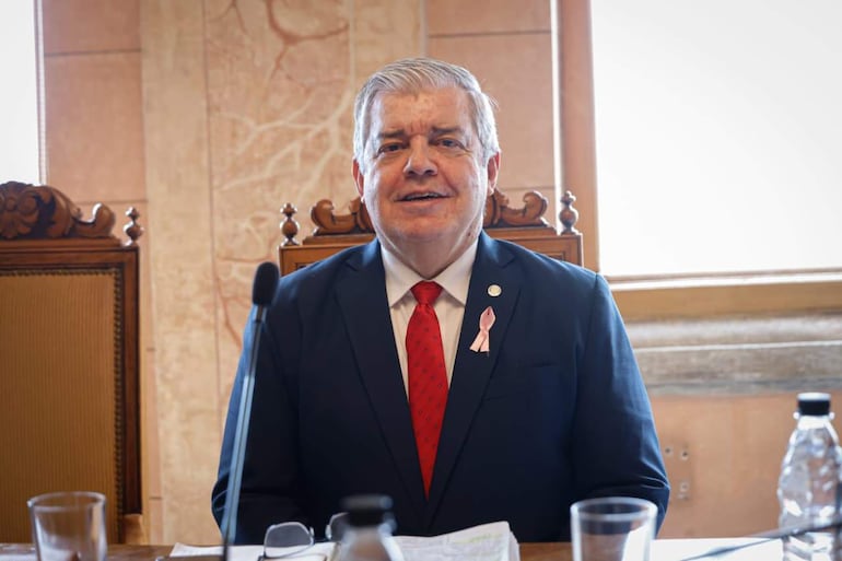 Enrique Riera Escudero sentado en mesa, con traje oscuro y corbata roja, en un entorno formal con documentos y micrófono.