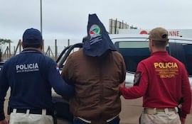 Dos policías en uniforme, uno azul y otro rojo, acompañan a un hombre con capucha, en un ambiente de detención con vehículos de fondo.