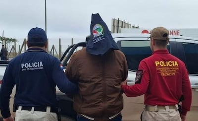 Dos policías en uniforme, uno azul y otro rojo, acompañan a un hombre con capucha, en un ambiente de detención con vehículos de fondo.