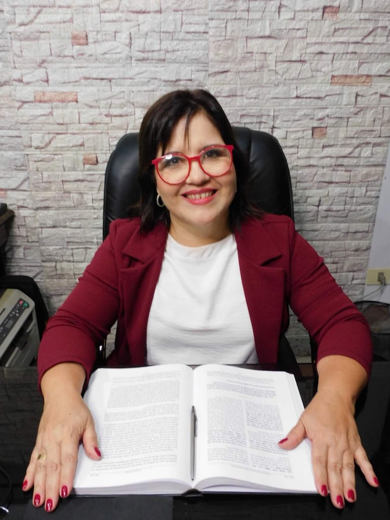 Mujer con gafas rojas y chaqueta burdeos sonríe, apoyando las manos sobre un libro en un entorno de oficina.