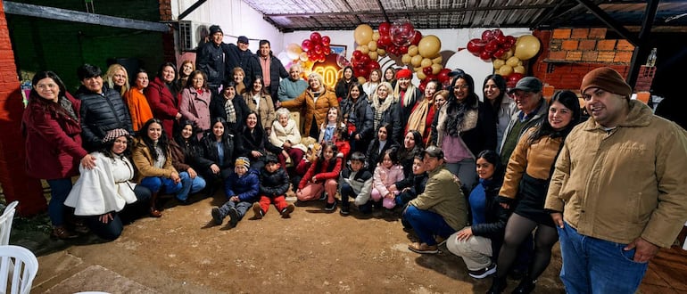 Doña Luciana en el centro, rodeada de familiares felices. Decoraciones festivas con globos rojos, dorados y cremosos.