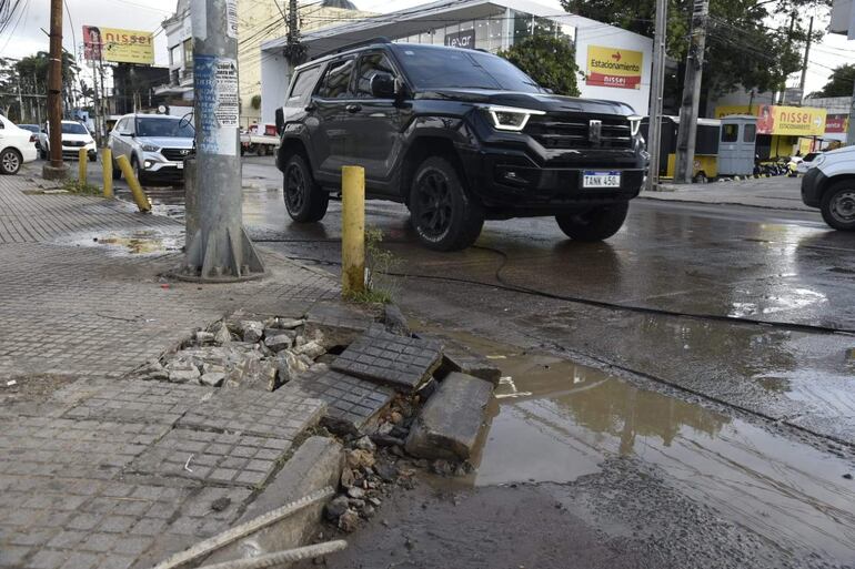 Entorno urbano con pavimento desgastado, postes de luz y vehículos, mientras un charco de agua estancada se forma en la calle.