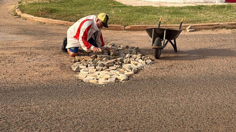 Hombre arrodillado con chaqueta blanca y sombrero amarillo colocando piedras en la calle. Carretilla vacía a su lado.