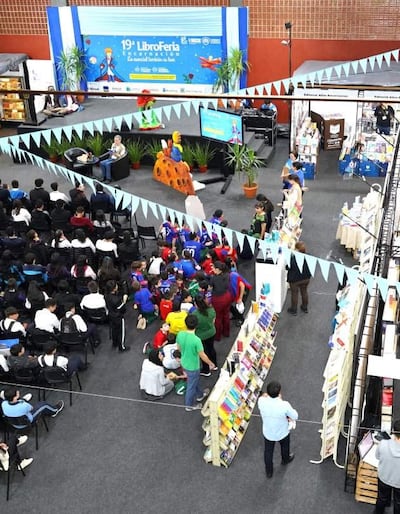 Grupo de jóvenes sentados en el suelo, escuchando a dos oradores en un escenario decorado con libros y banderines de colores.