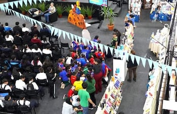 Grupo de jóvenes sentados en el suelo, escuchando a dos oradores en un escenario decorado con libros y banderines de colores.