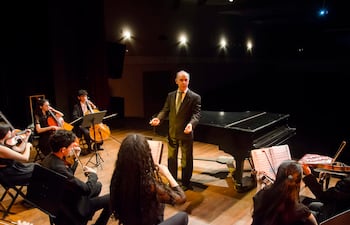 Músicos jóvenes en concierto, vestimenta formal, director al frente con traje negro y gestos enérgicos, piano negro de fondo.