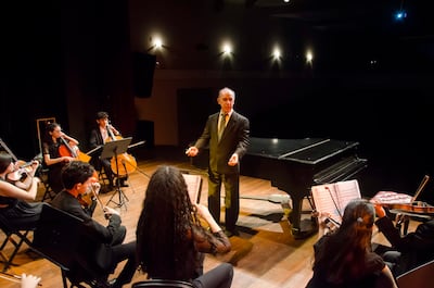 Músicos jóvenes en concierto, vestimenta formal, director al frente con traje negro y gestos enérgicos, piano negro de fondo.