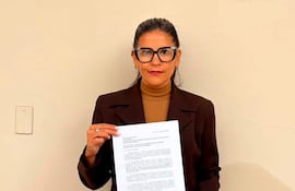 Noelia Díaz de pie, sosteniendo un documento con abrigo oscuro y pantalones claros, mirando hacia la cámara en un ambiente interior.