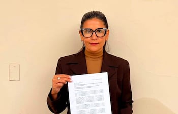 Noelia Díaz de pie, sosteniendo un documento con abrigo oscuro y pantalones claros, mirando hacia la cámara en un ambiente interior.