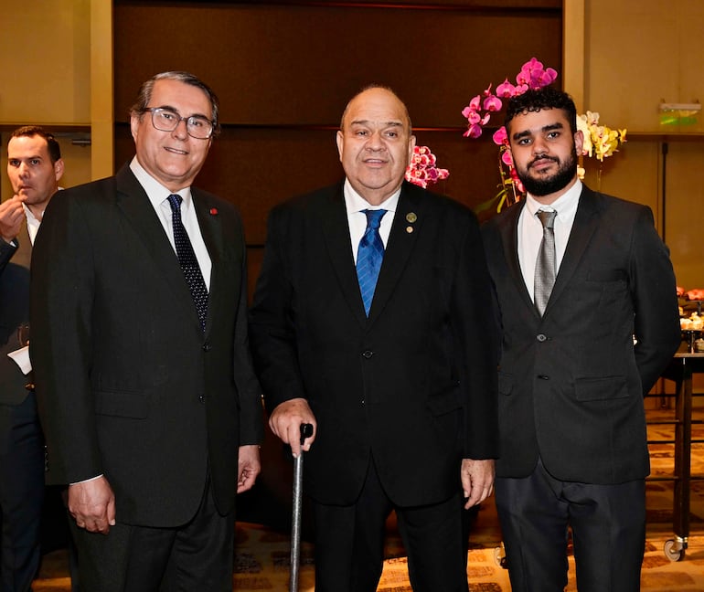 Tres hombres con trajes oscuros en ambiente formal, uno con bastón, posando con sonrisas en decoración floral.
