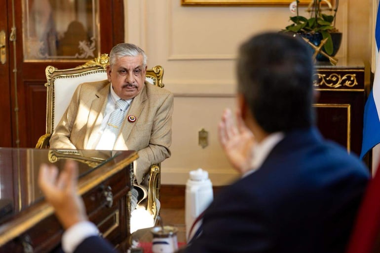 Isaías Ricardo Fretes sentado en silla ornamentada, vestido con chaqueta beige y corbata clara, escucha a un hombre de pie gesticulando.