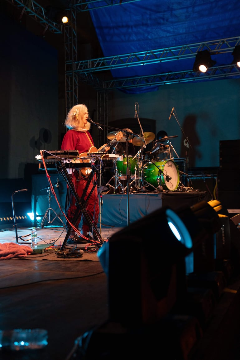 Mujer rubia en el escenario con guitarra, cantando, rodeada de instrumentos de banda en un evento al aire libre.