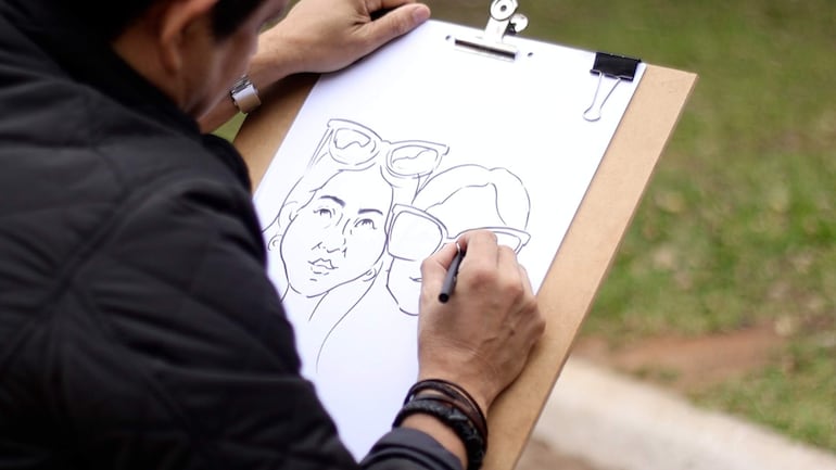 Artista en abrigo negro dibujando con pluma en un clipboard, rodeado de césped y un camino.