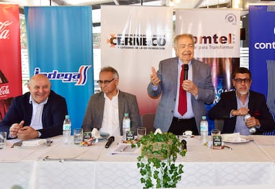 Hombre en el centro gesticula mientras habla, flanqueado por otros tres hombres, en un salón con banners publicitarios.