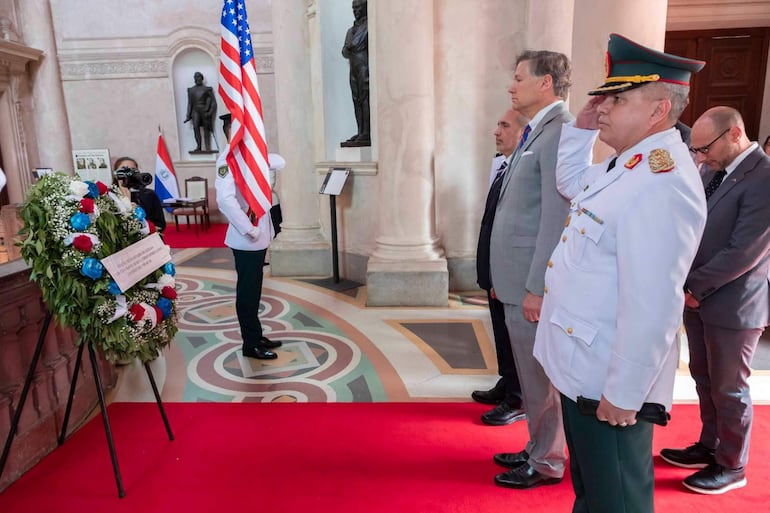 Robert Alter y Víctor Verdún en trajes oscuros, mientras un oficial militar saluda con uniforme blanco, rodeados de columnas y ofrendas.