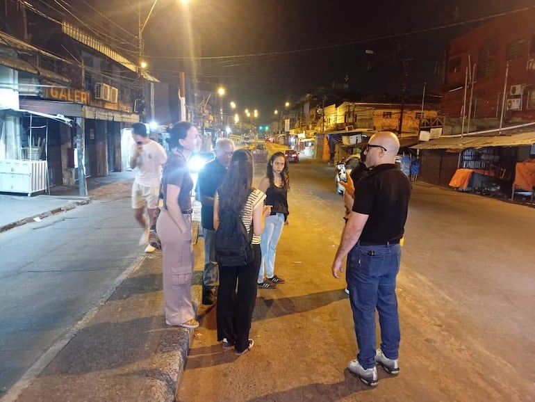 Hombre con gafas sostiene un teléfono, mientras mujer con cabello rizado escucha. Ambiente urbano de noche con luces y tiendas al fondo.
