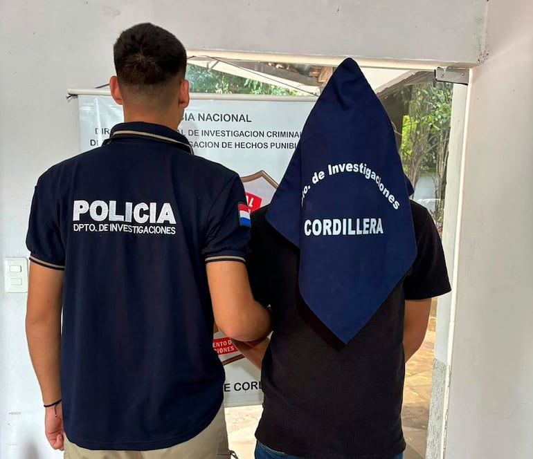 Agente de policía en camiseta negra y persona con paño oscuro en la cabeza, en un entorno interior con cartel policial visible.