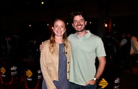 Mujer rubia en vestido azul y chaqueta clara sonríe junto a hombre de camiseta verde en ambiente decorado con llantas.