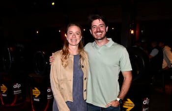 Mujer rubia en vestido azul y chaqueta clara sonríe junto a hombre de camiseta verde en ambiente decorado con llantas.